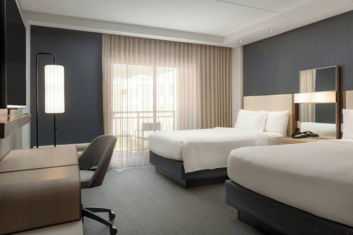 Ein Hotelzimmer mit zwei Betten, einem Schreibtisch, einem Stuhl, einer Lampe und einem großen Fenster mit Vorhängen.
