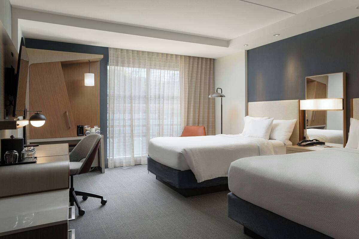 Ein Hotelzimmer mit Schreibtisch, zwei Betten, einem Sessel, einer Schreibtischlampe, einem Nachttisch und einem großen Spiegel.