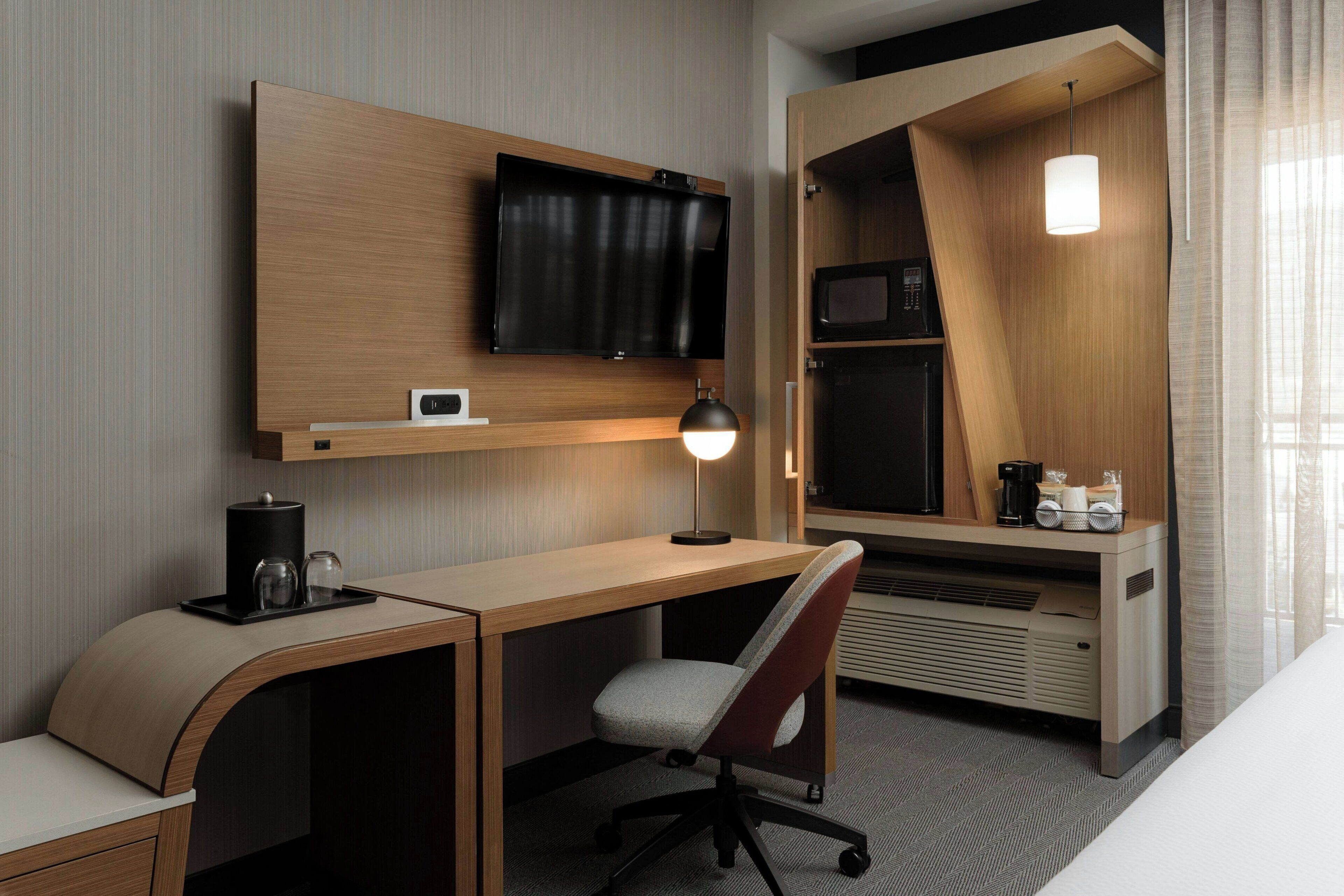 A hotel room with a wooden desk, a television mounted on the wall, a microwave, a coffee maker, and a bed with a white comforter.