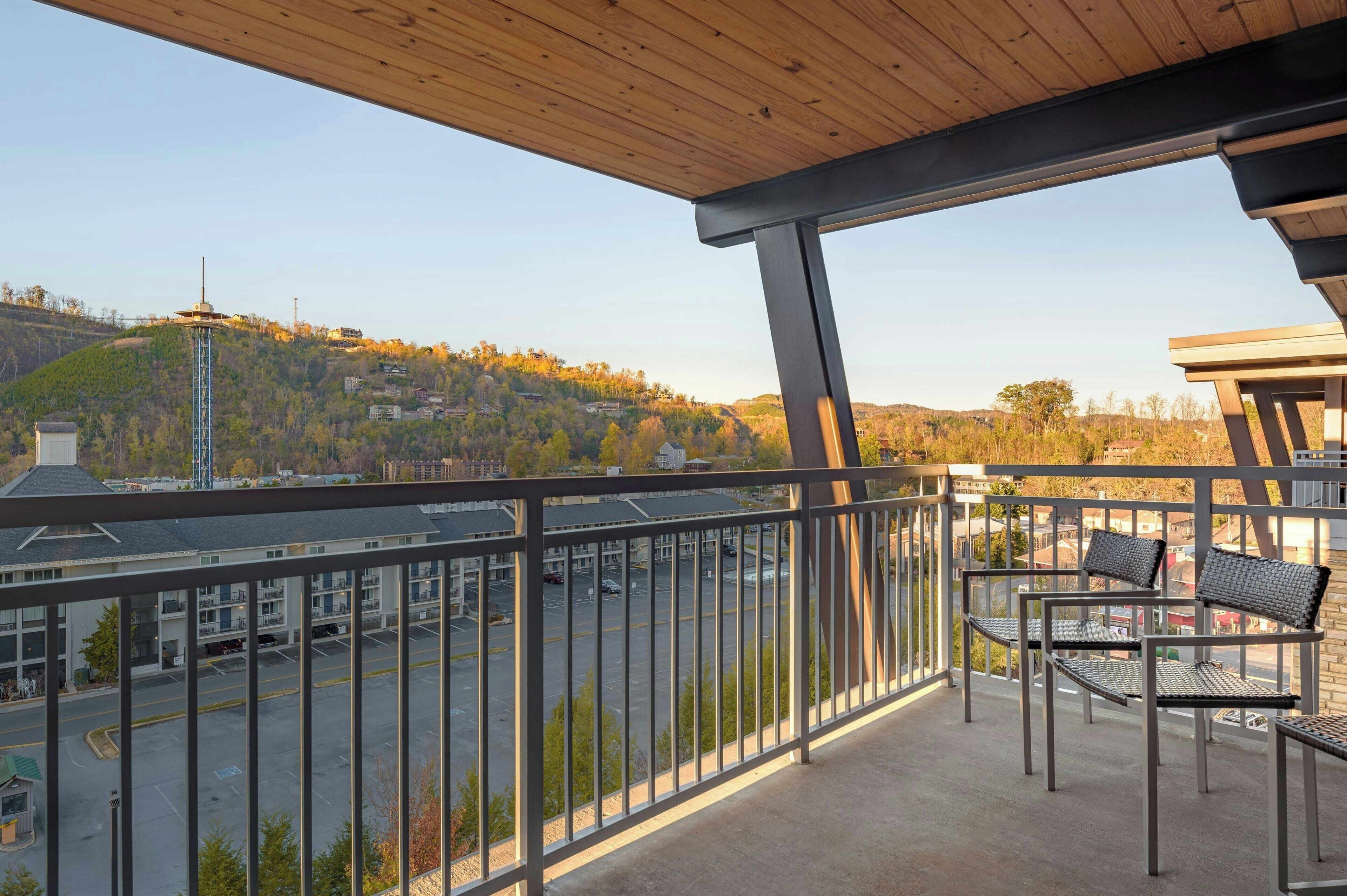 A balcony with a view of a parking lot and buildings, a tower, and a hilly landscape.