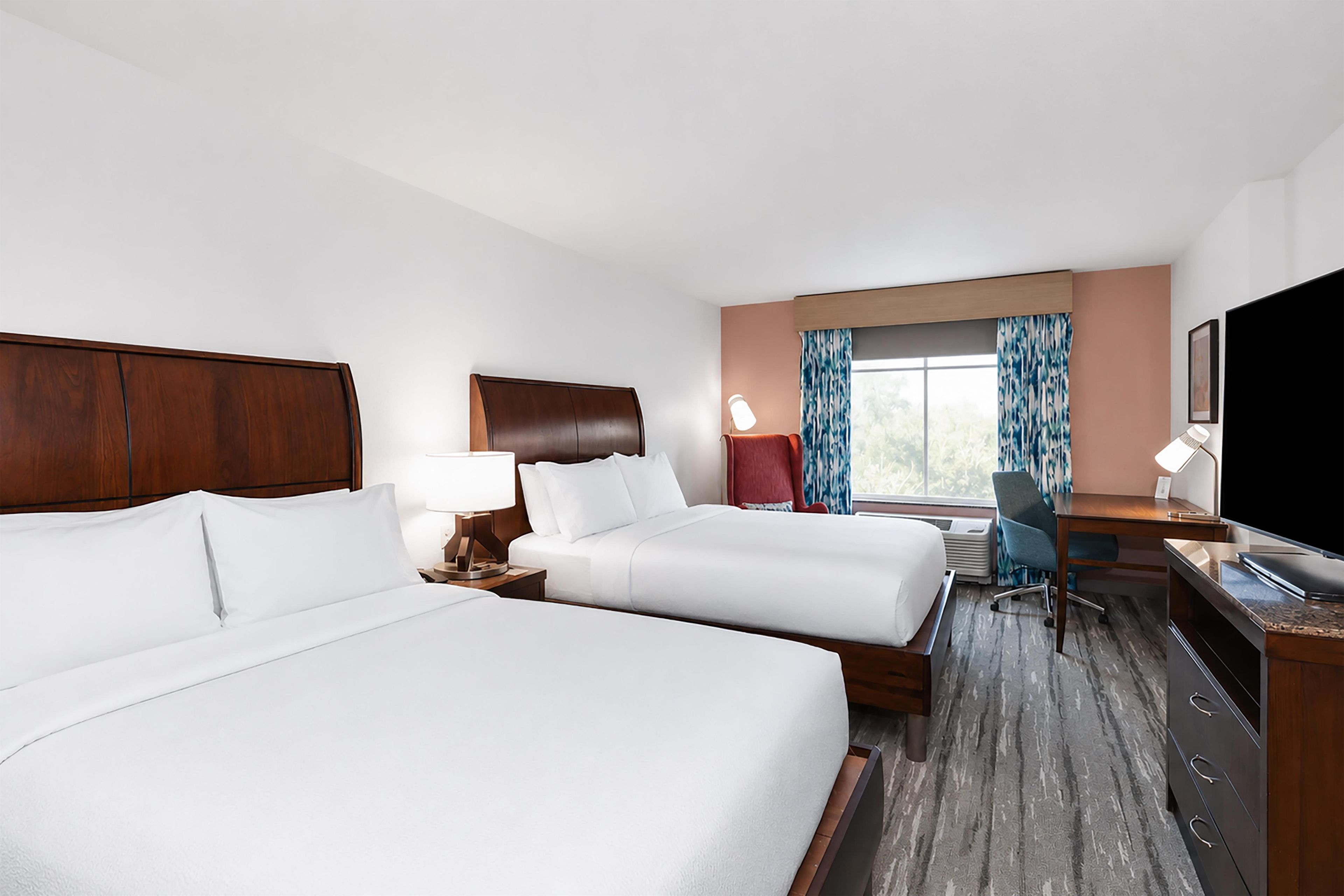A hotel room with two beds, a desk, a chair, a TV, and a window with curtains.