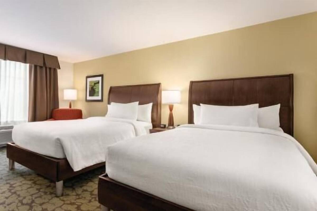 A hotel room with two beds, a red chair, a lamp, and a framed picture on the wall.