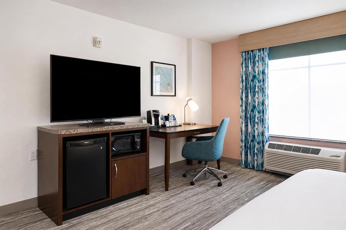 A hotel room with a flat-screen TV, a desk with a chair, a microwave, a small refrigerator, a bedside table, and a large window with curtains.