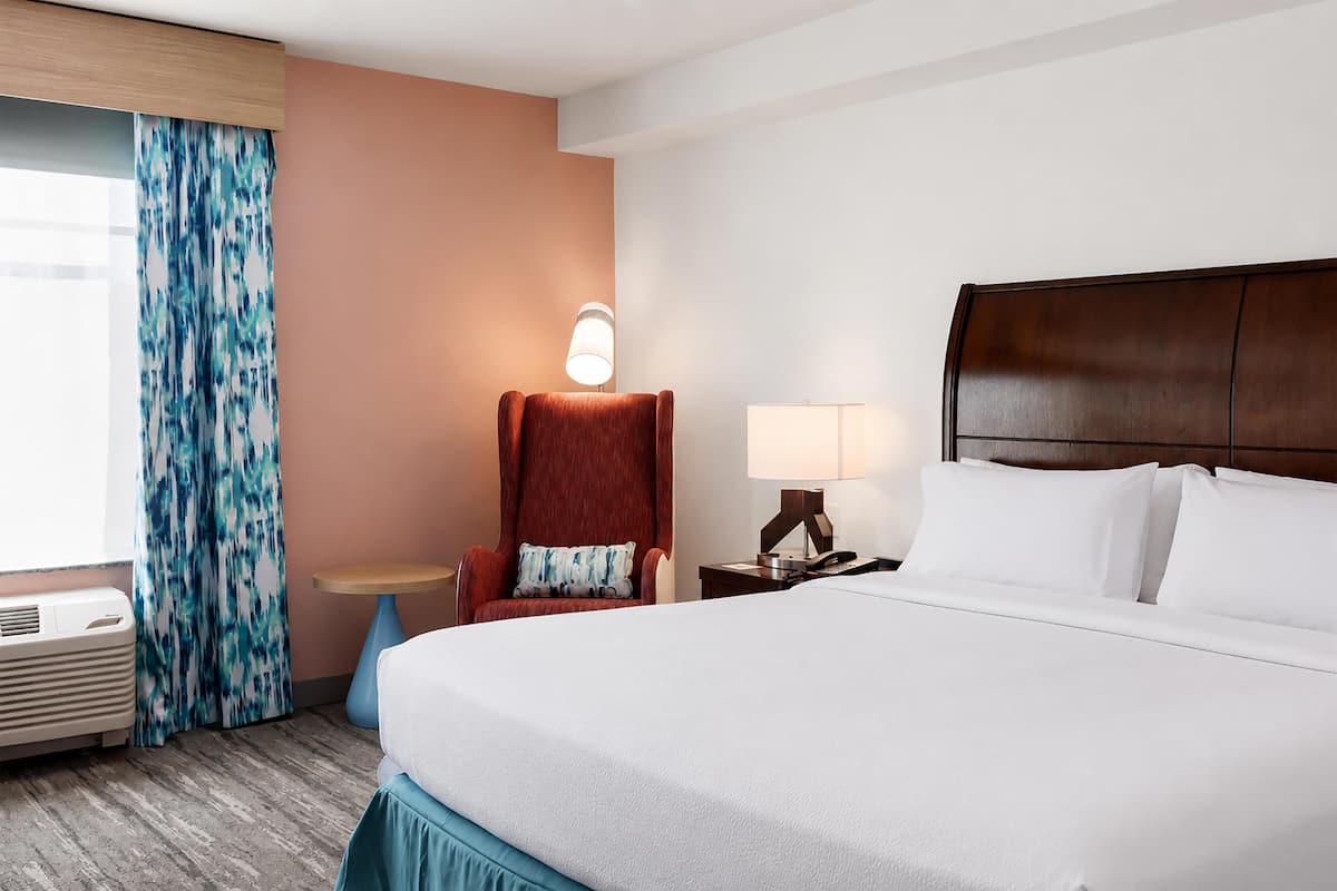 A hotel room with a bed, a chair, a table, a lamp, and a window with curtains.