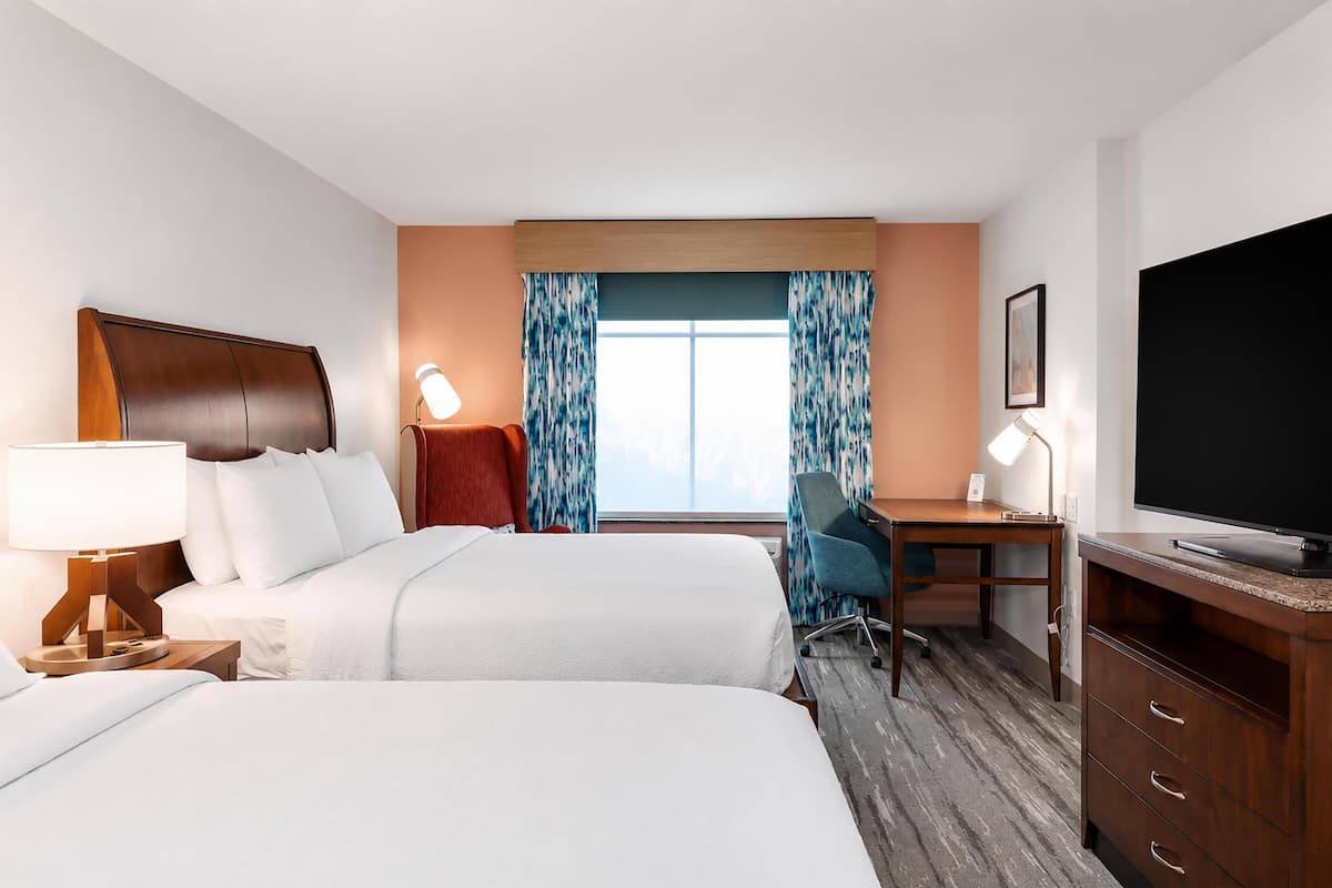 A hotel room with two beds, a desk, a TV, and a window with curtains.