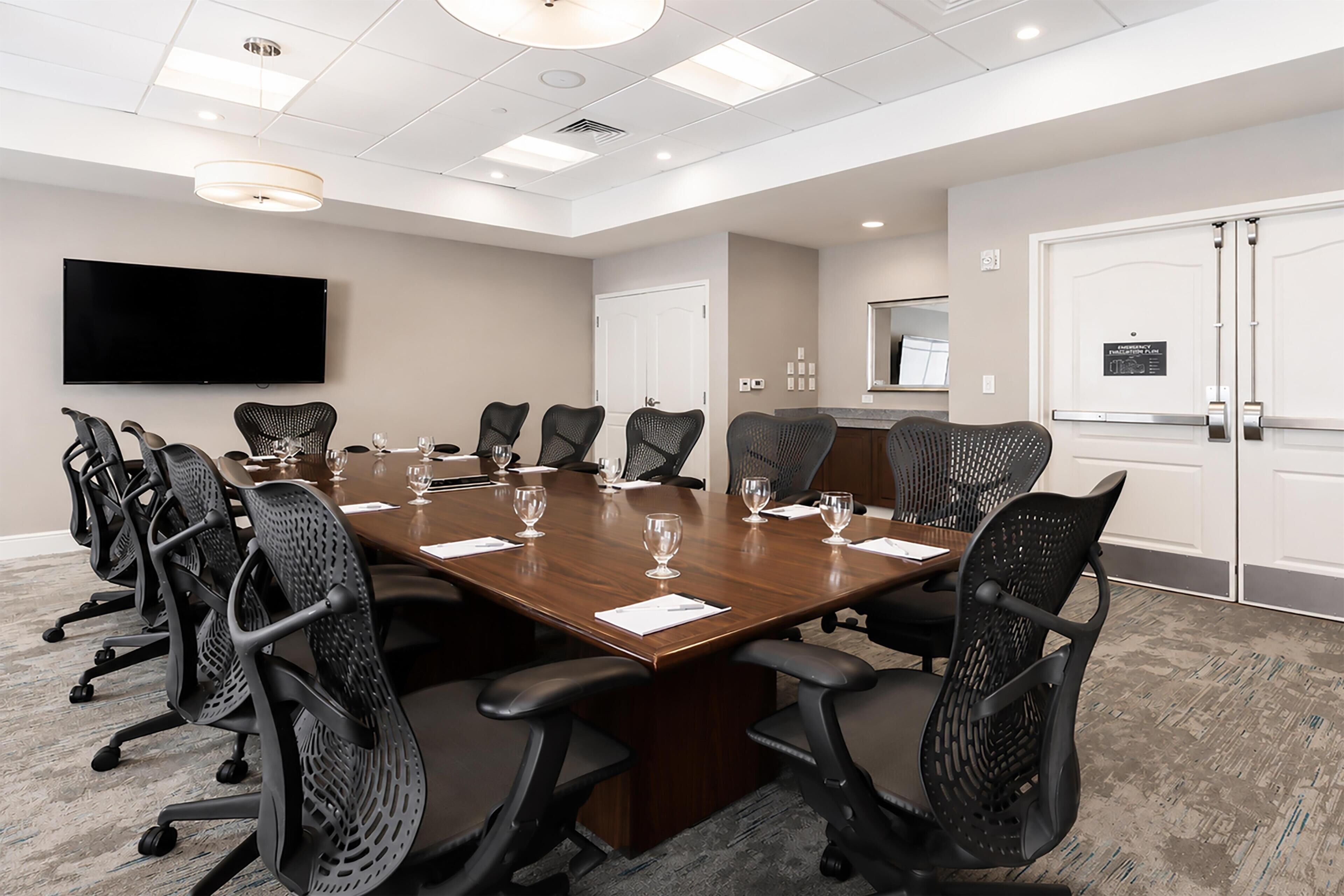 A conference room with a long table, chairs, a flat-screen TV, and a door with a sign.