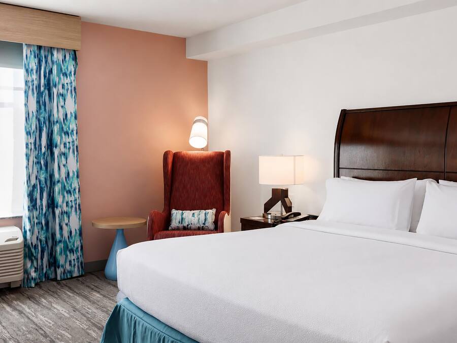 A hotel room with a bed, a chair, a table, a lamp, and a window with curtains.