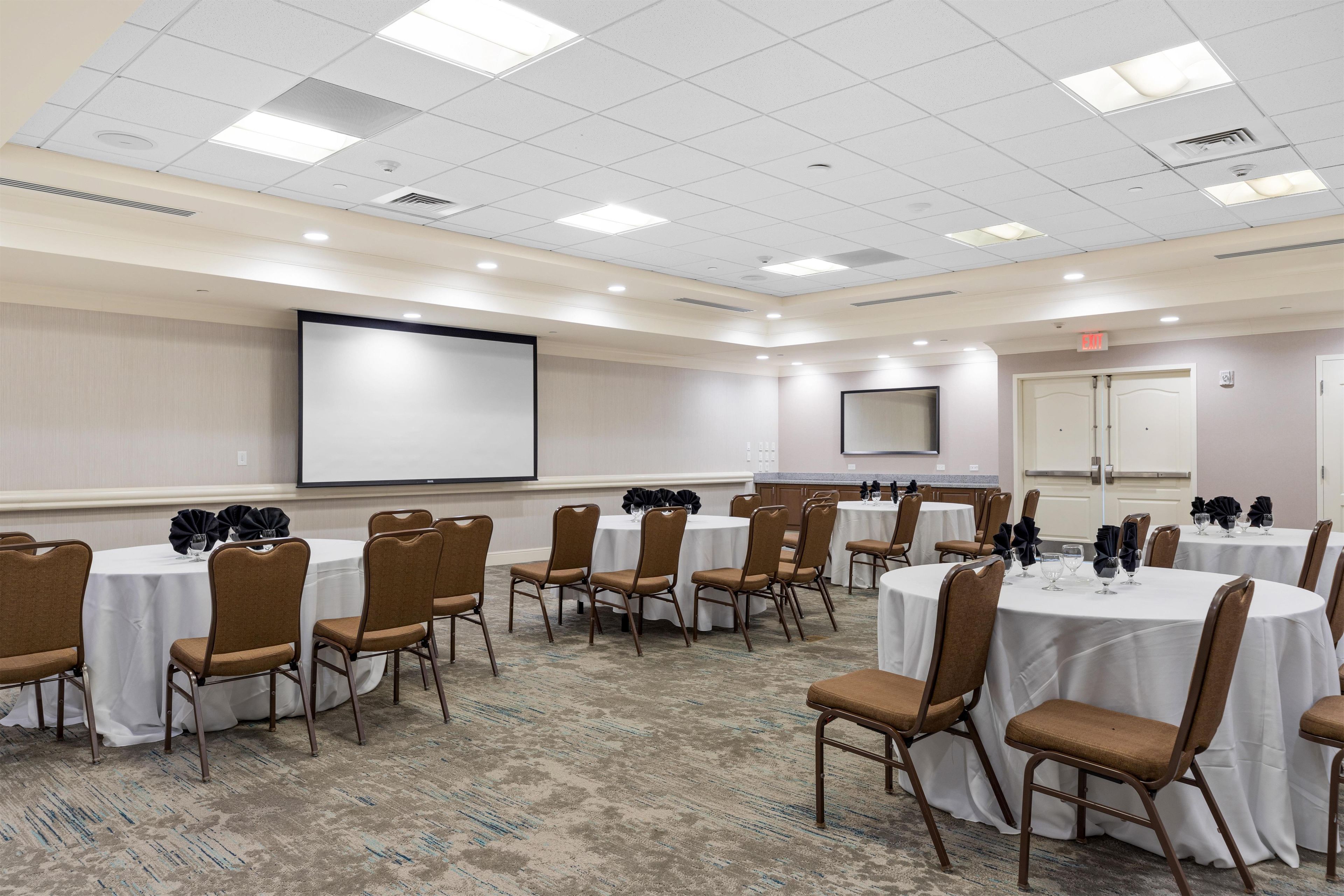 A conference room with multiple tables and chairs, a projector screen, and an exit sign.