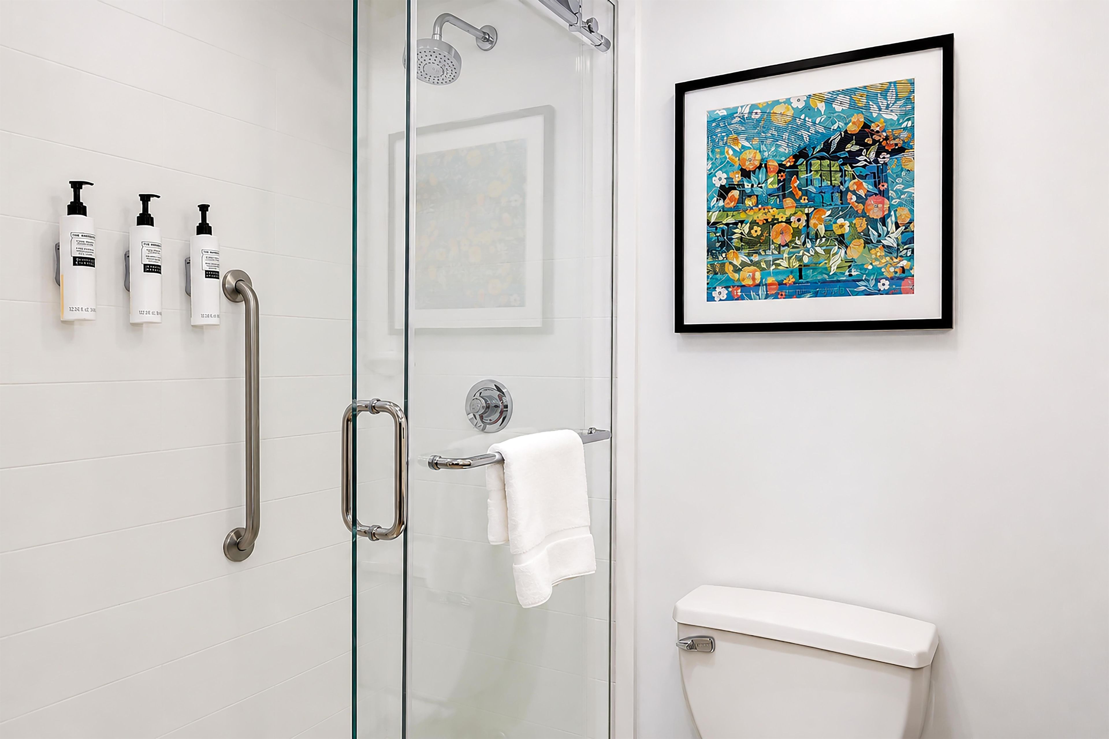 Un baño con cabina de ducha de vidrio, un inodoro blanco, una obra de arte enmarcada en la pared y tres botellas de champú.