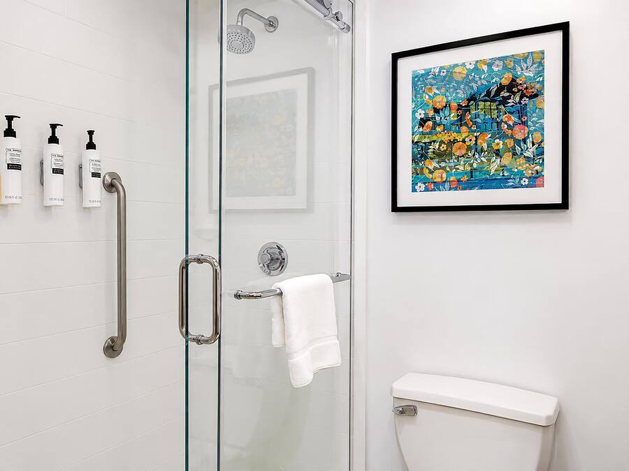 A bathroom with a glass shower enclosure, a white toilet, a framed artwork on the wall, and three bottles of shampoo.