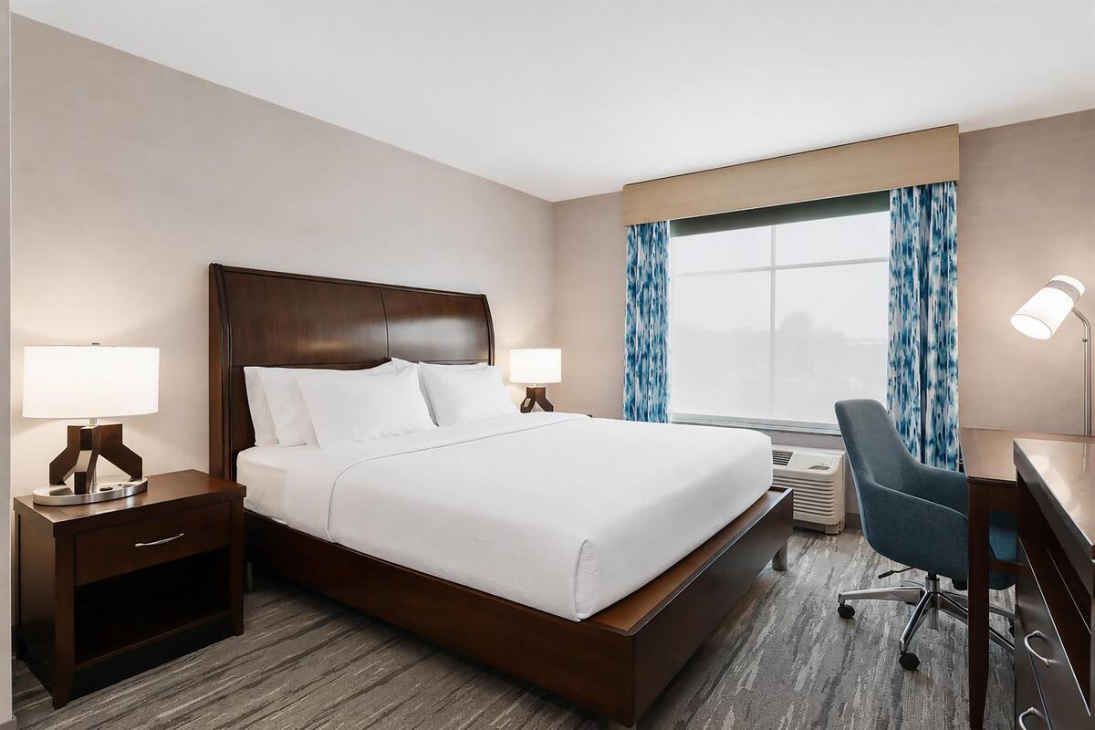 A hotel room with a large bed, bedside tables with lamps, a desk with a chair, and a window with curtains.