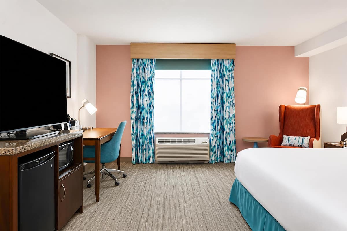 A hotel room with a bed, desk, television, air conditioning unit, and a chair.