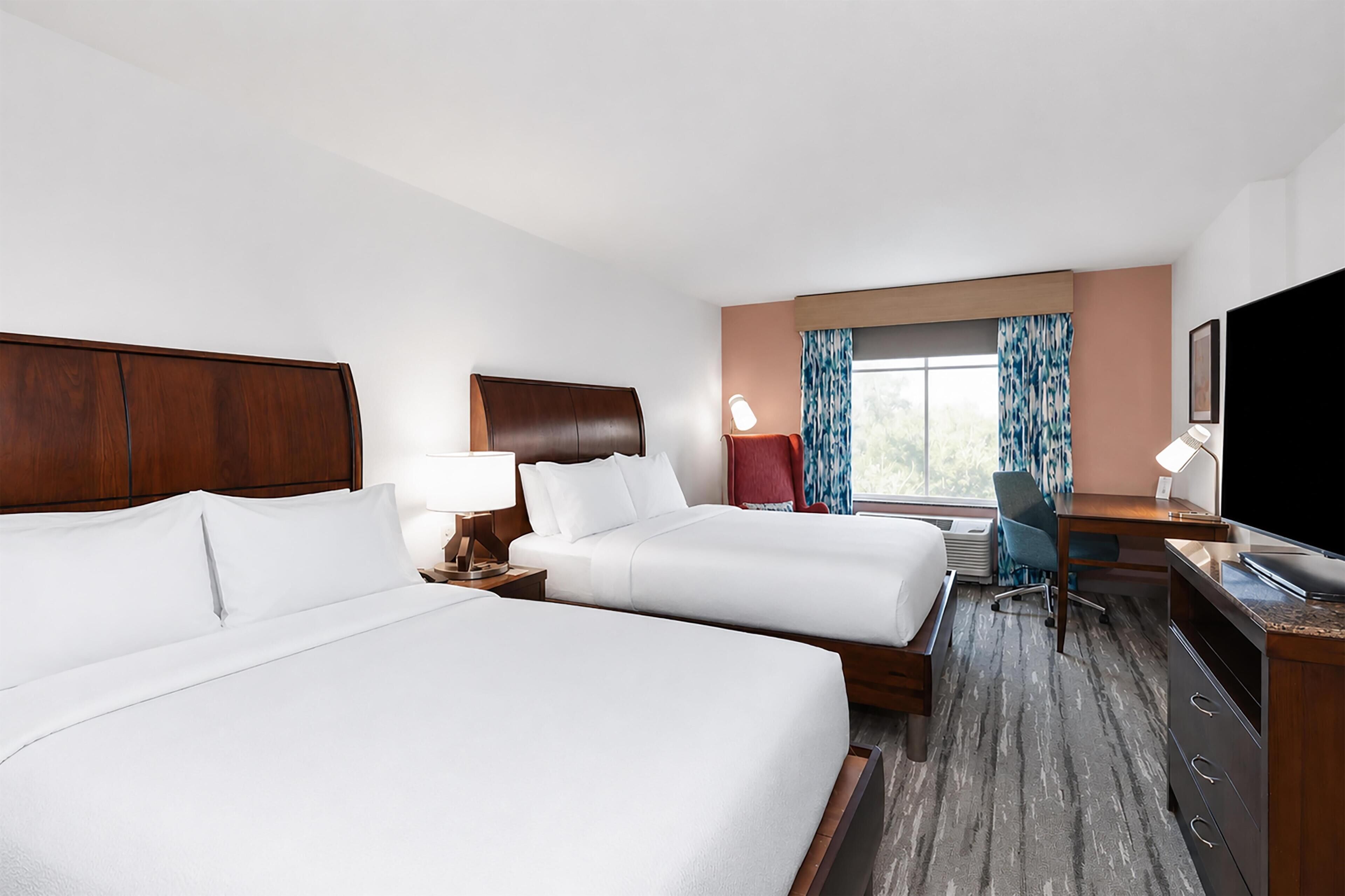 A hotel room with two beds, a desk, a chair, a TV, and a window with curtains.