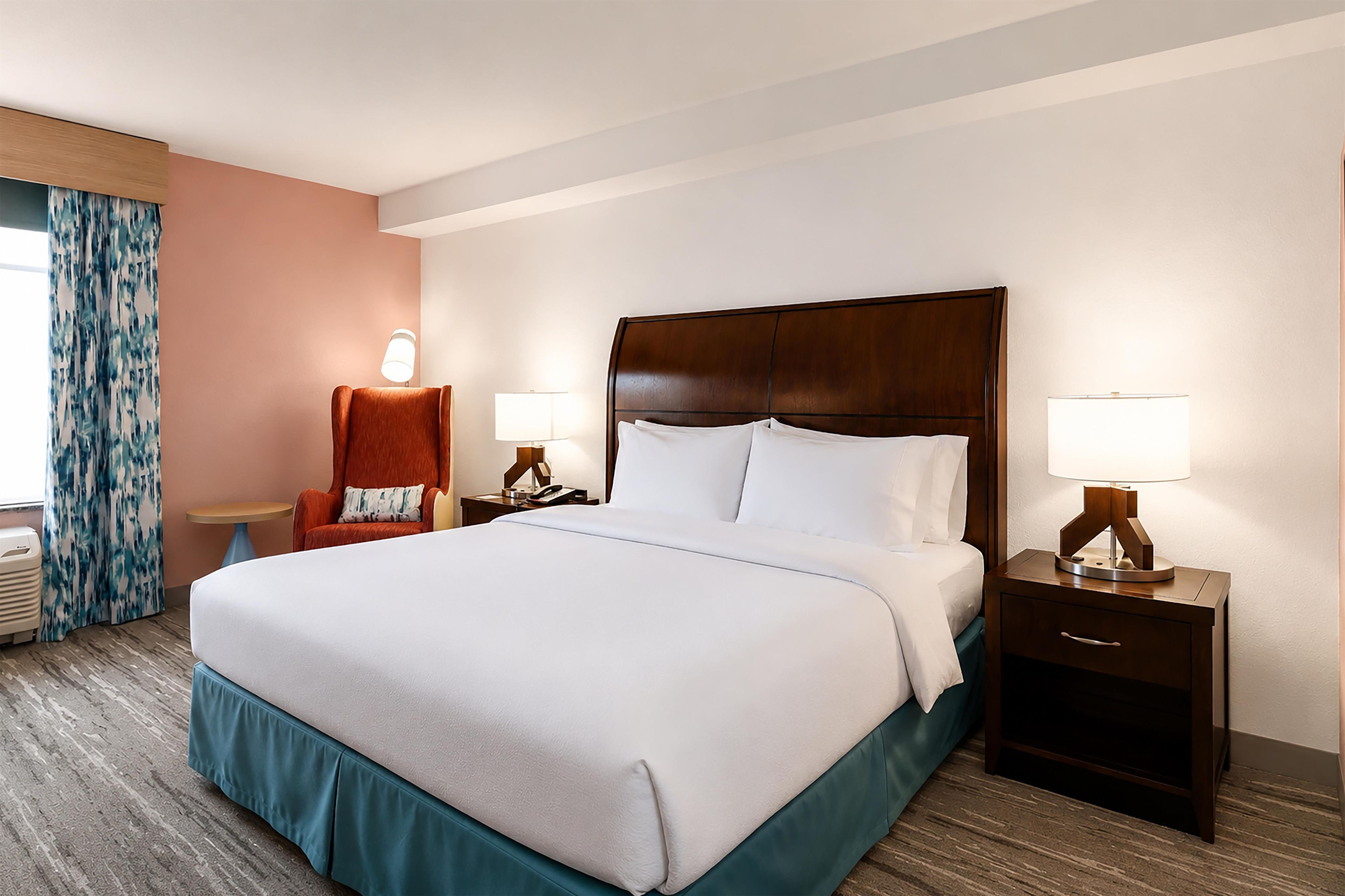A hotel room with a large bed, two bedside tables with lamps, a chair, and a window with curtains.