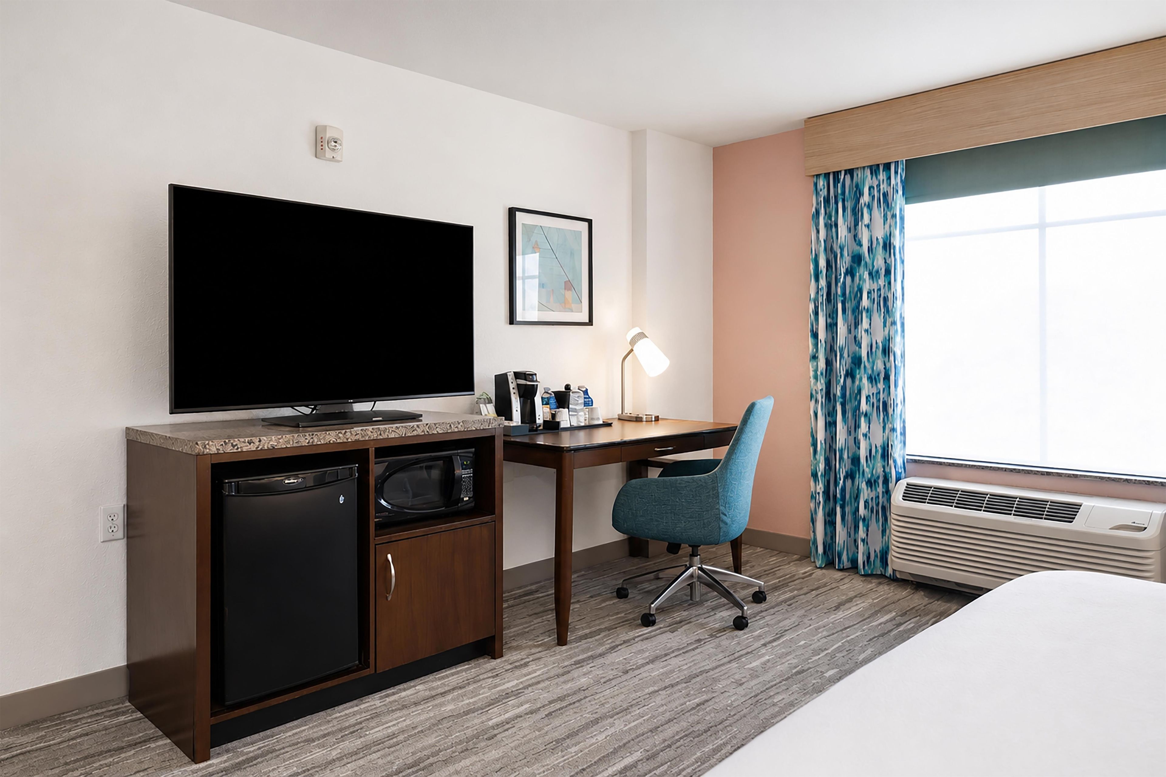A hotel room with a flat-screen TV, a desk with a chair, a microwave, a small refrigerator, a bedside table, and a large window with curtains.