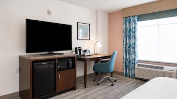 A hotel room with a flat-screen TV, a desk with a chair, a microwave, a small refrigerator, a bedside table, and a large window with curtains.