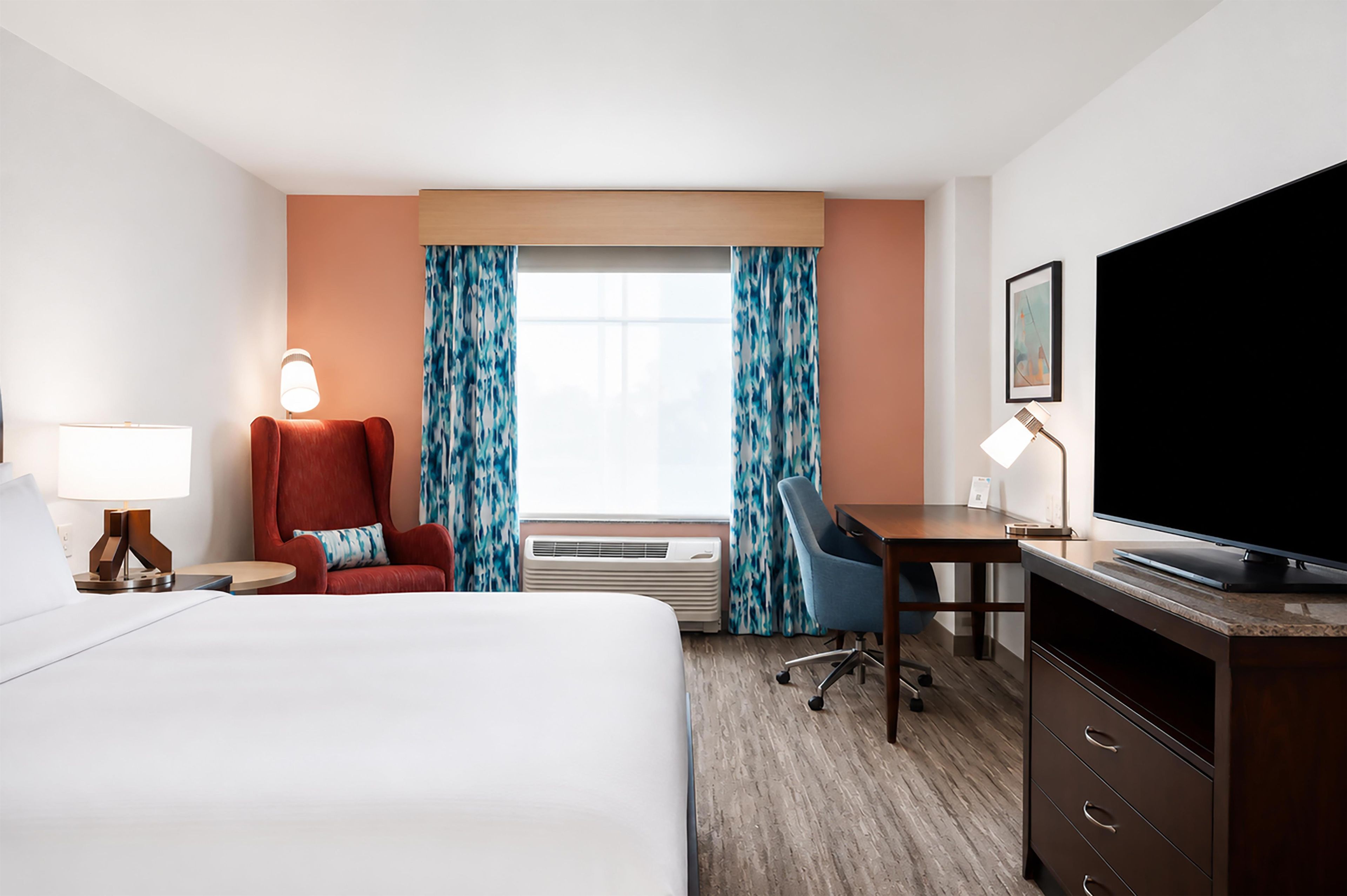 A hotel room with a bed, desk, chair, television, and air conditioning unit.