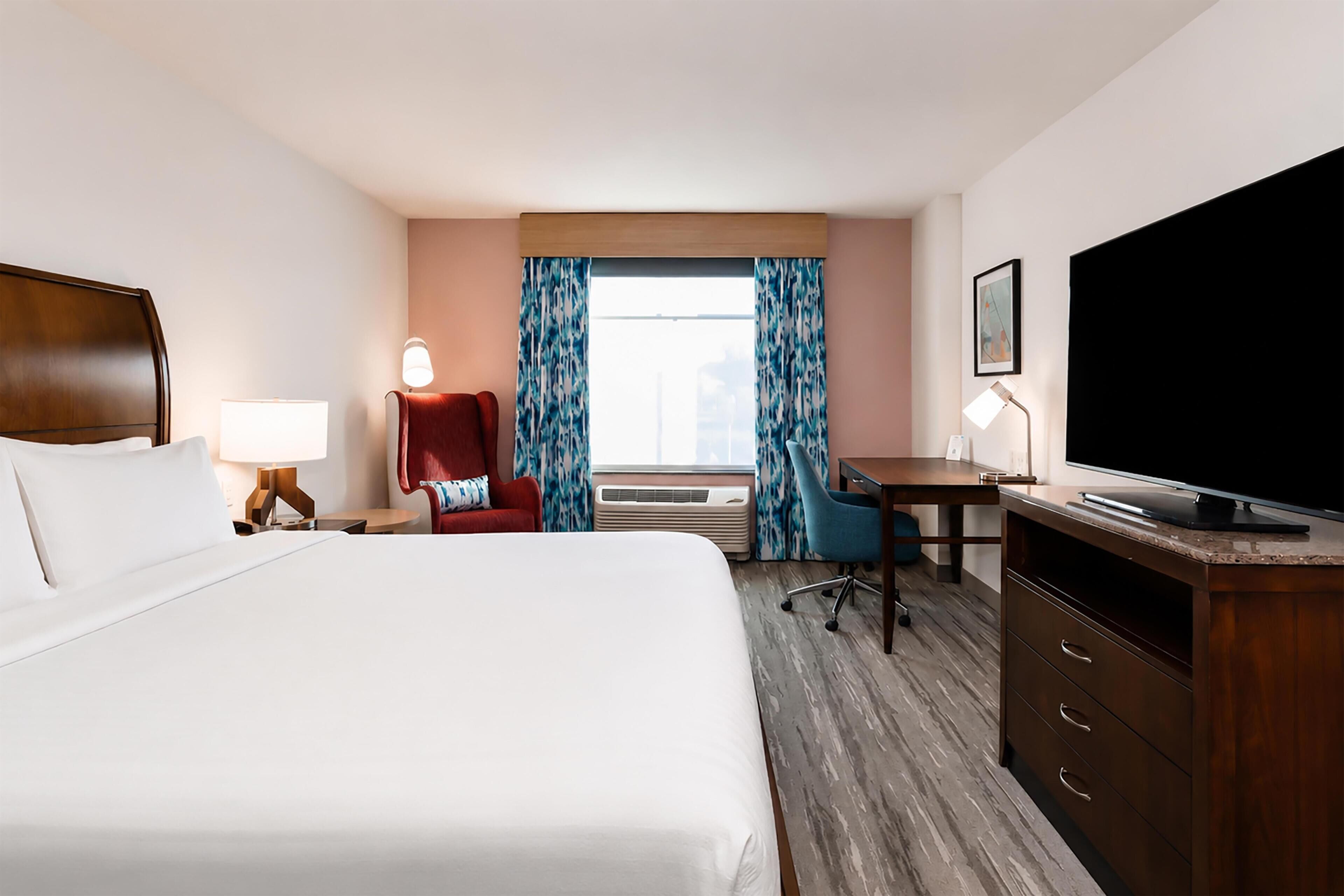 A hotel room with a bed, desk, TV, chair, lamp, and air conditioning unit.