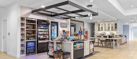 A modern hotel lobby with a central bar area, refrigerated beverage dispensers, and a seating area with tables and chairs.