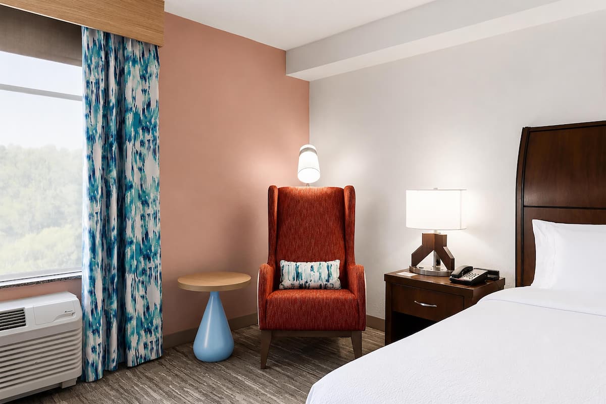 A hotel room with a bed, a chair, a small table, a lamp, a phone, and a window with curtains.