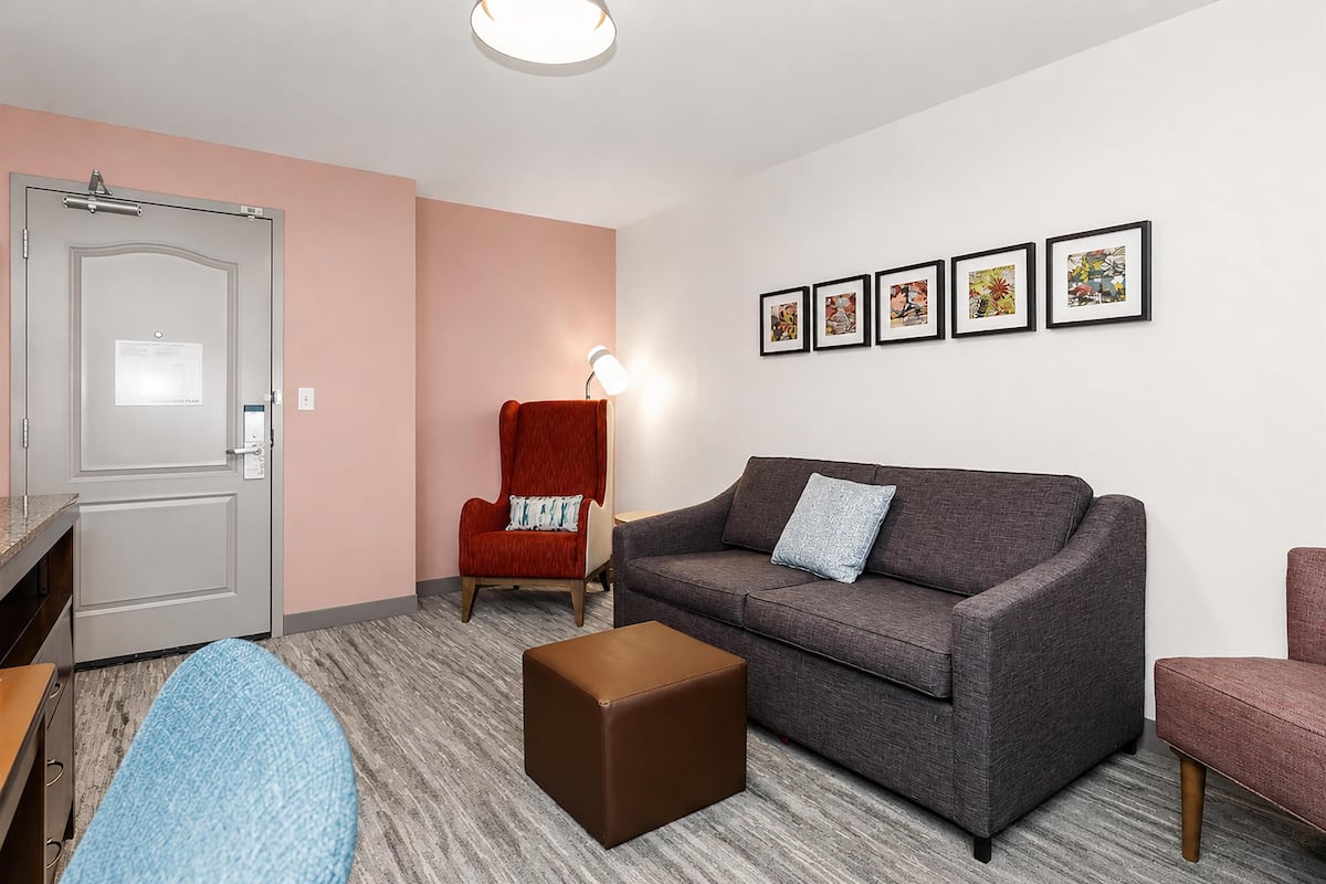 A living room with a grey sofa, a red armchair, a brown ottoman, and a door with a light switch.