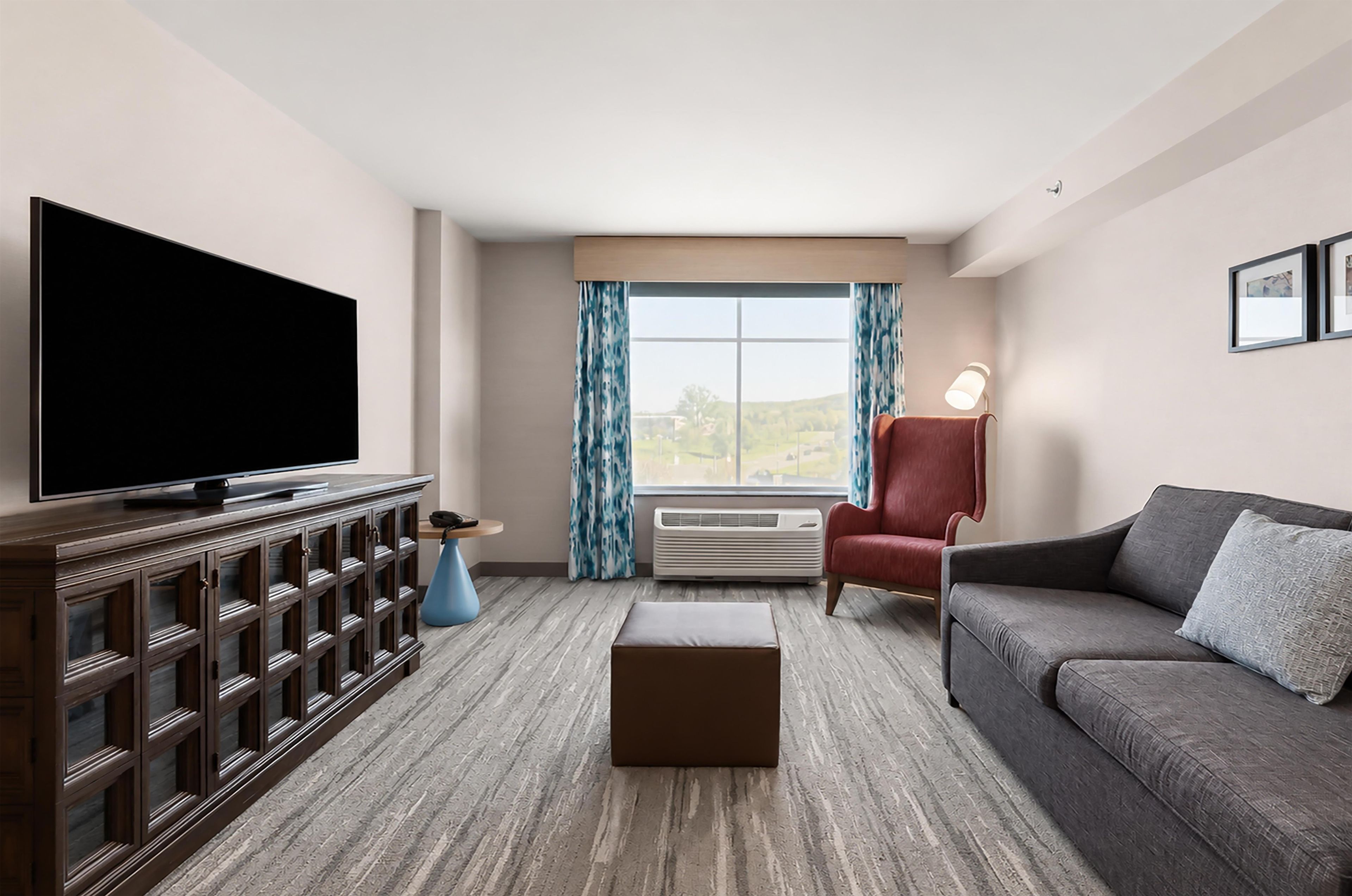 A modern living room with a flat-screen TV, a red armchair, a grey sofa, a wooden entertainment unit, and a large window with curtains.