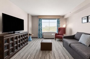 A modern living room with a flat-screen TV, a red armchair, a grey sofa, a wooden entertainment unit, and a large window with curtains.
