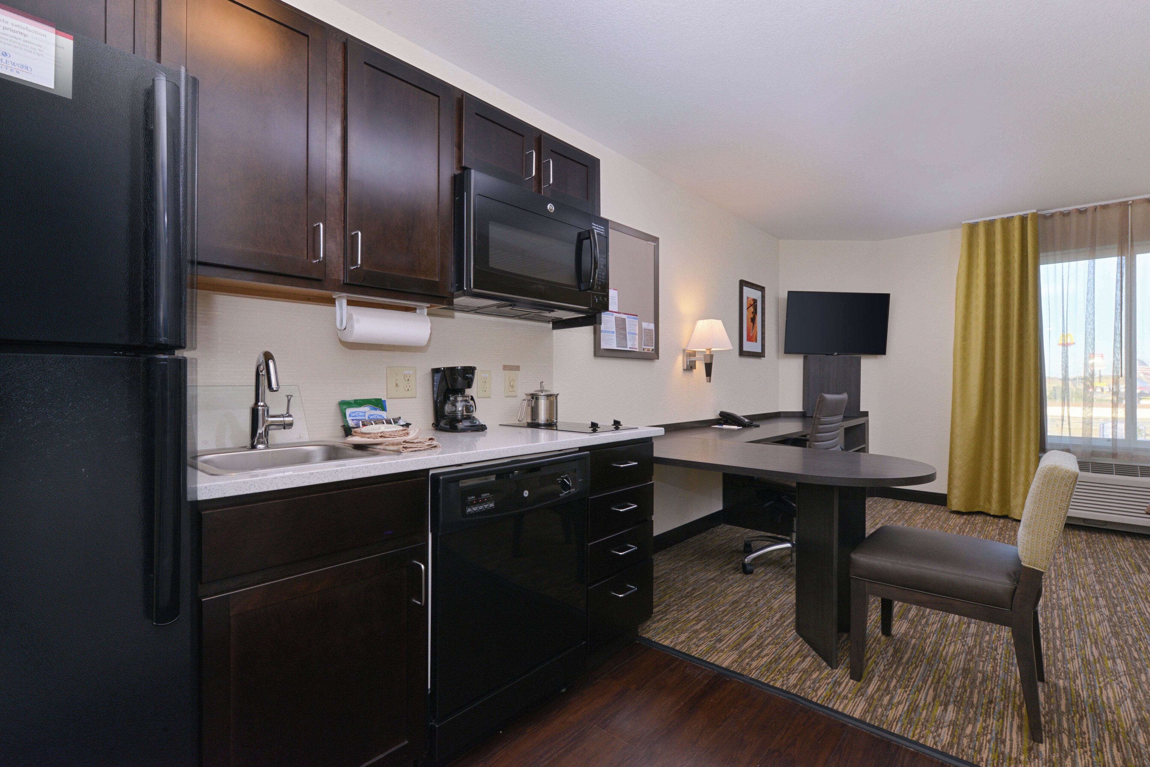 A compact hotel kitchen with a refrigerator, microwave, coffee maker, and a small dining area with a chair and table.