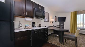 A compact hotel kitchen with a refrigerator, microwave, coffee maker, and a small dining area with a chair and table.