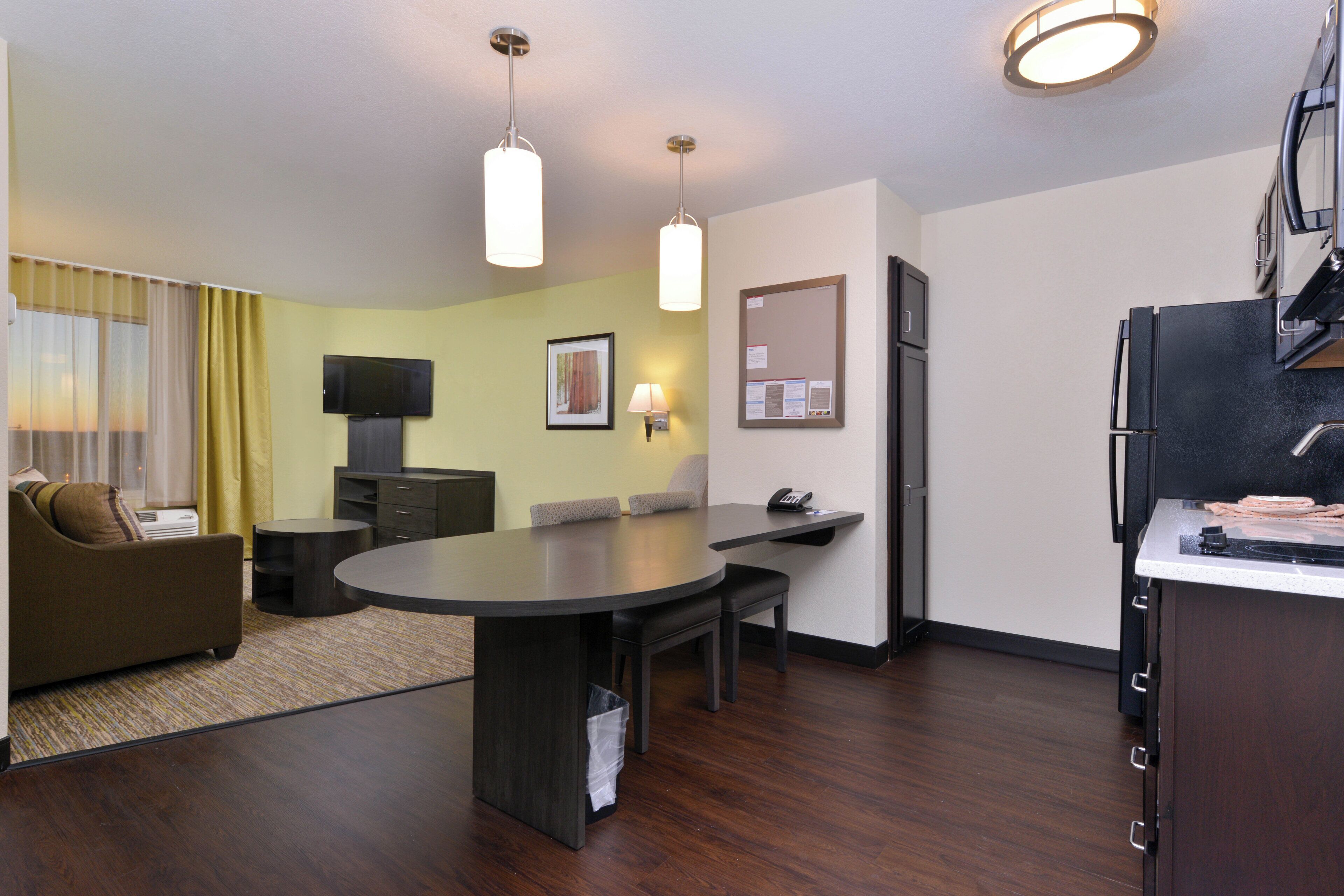 A modern hotel room with a dining area, a flat-screen TV, a sofa, and a kitchen area with a refrigerator.