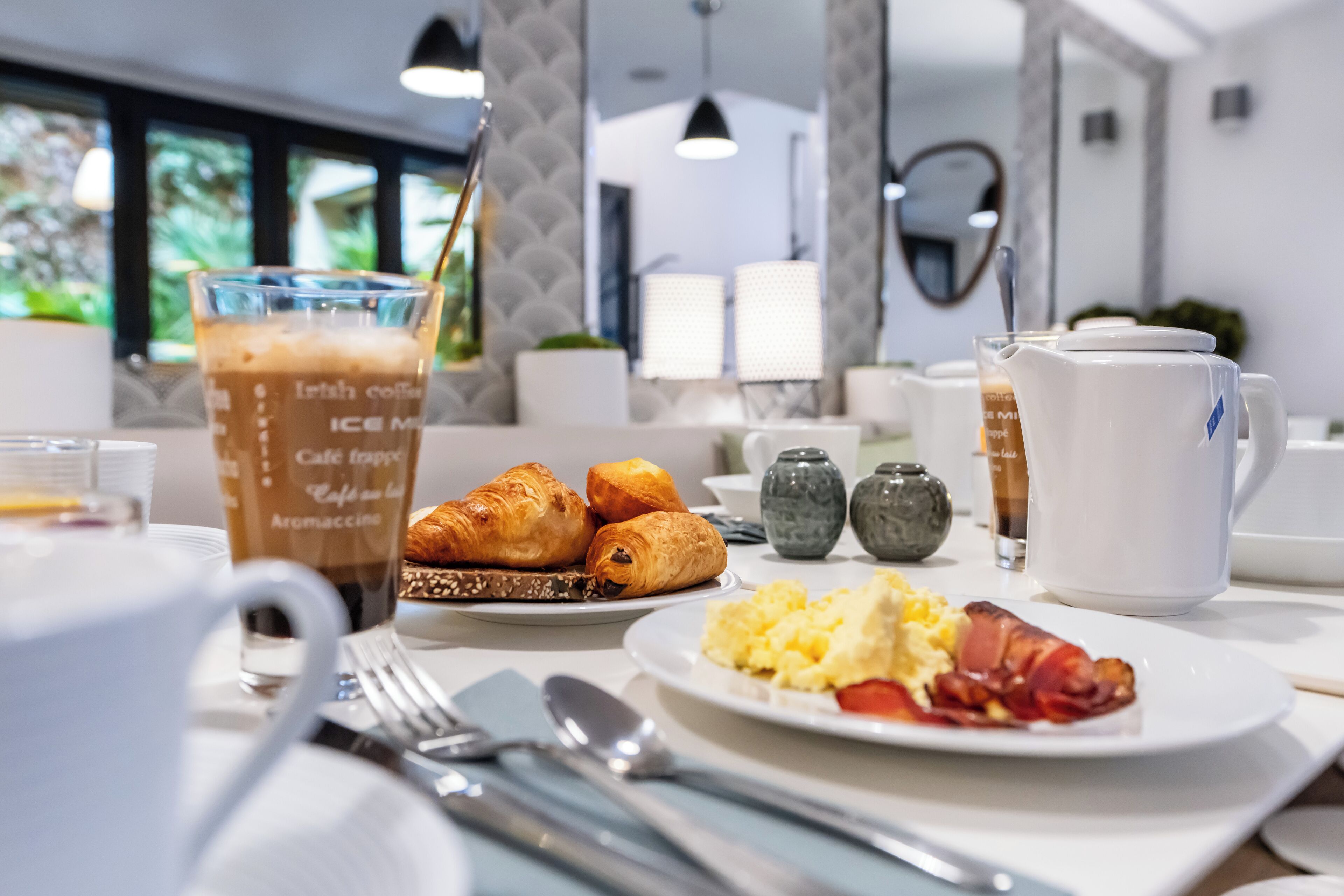 A breakfast table with croissants, scrambled eggs, bacon, and a coffee drink.