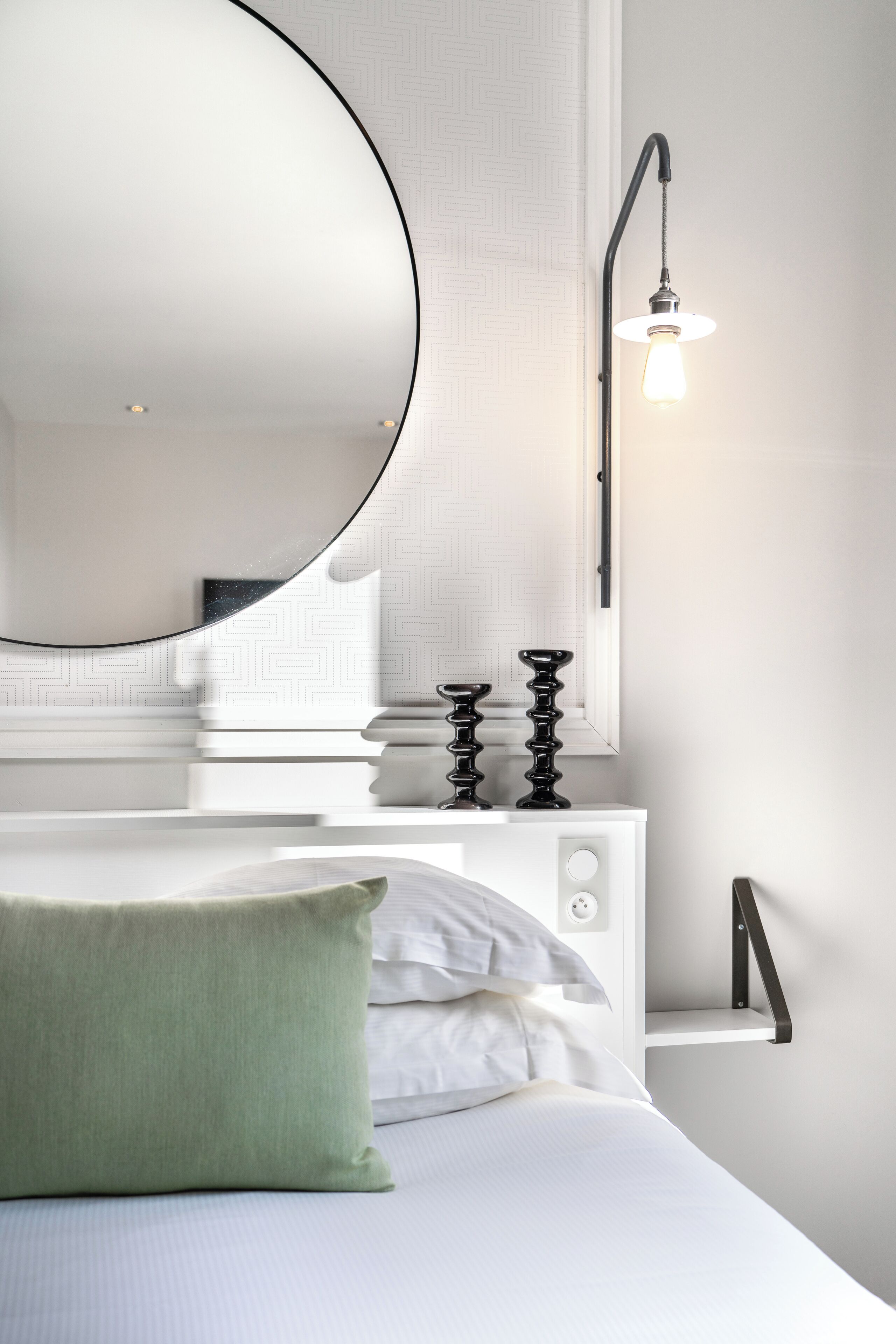 Quarto moderno com um espelho redondo grande, uma mesa de cabeceira com abajur e uma cama com travesseiro verde.