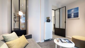 Un salon moderne avec un canapé gris, une petite table blanche et un pouf beige. Un grand miroir reflète un couloir et une porte.