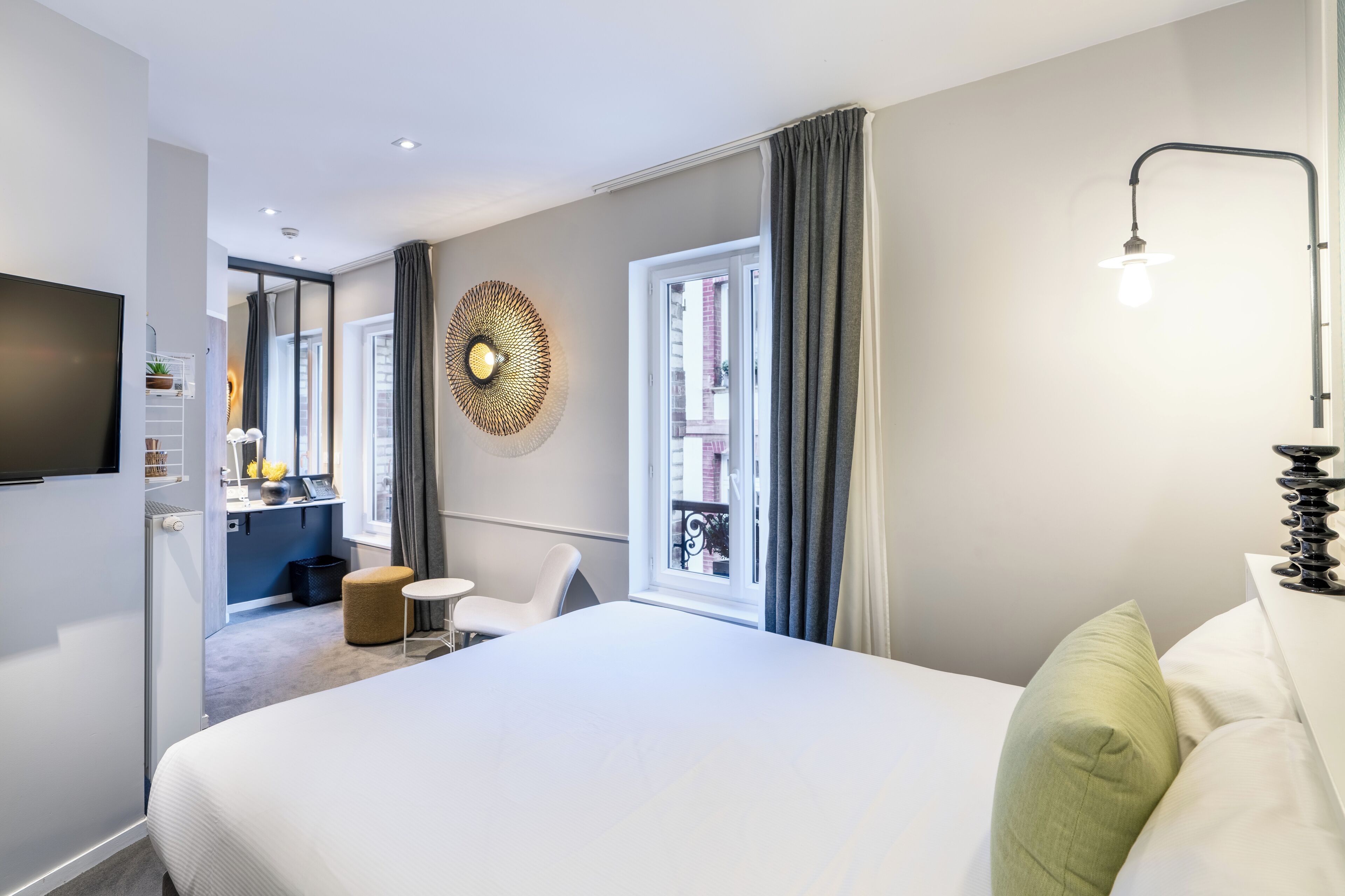 A modern hotel room with a large bed, a small desk, a chair, a TV, and a window with curtains.