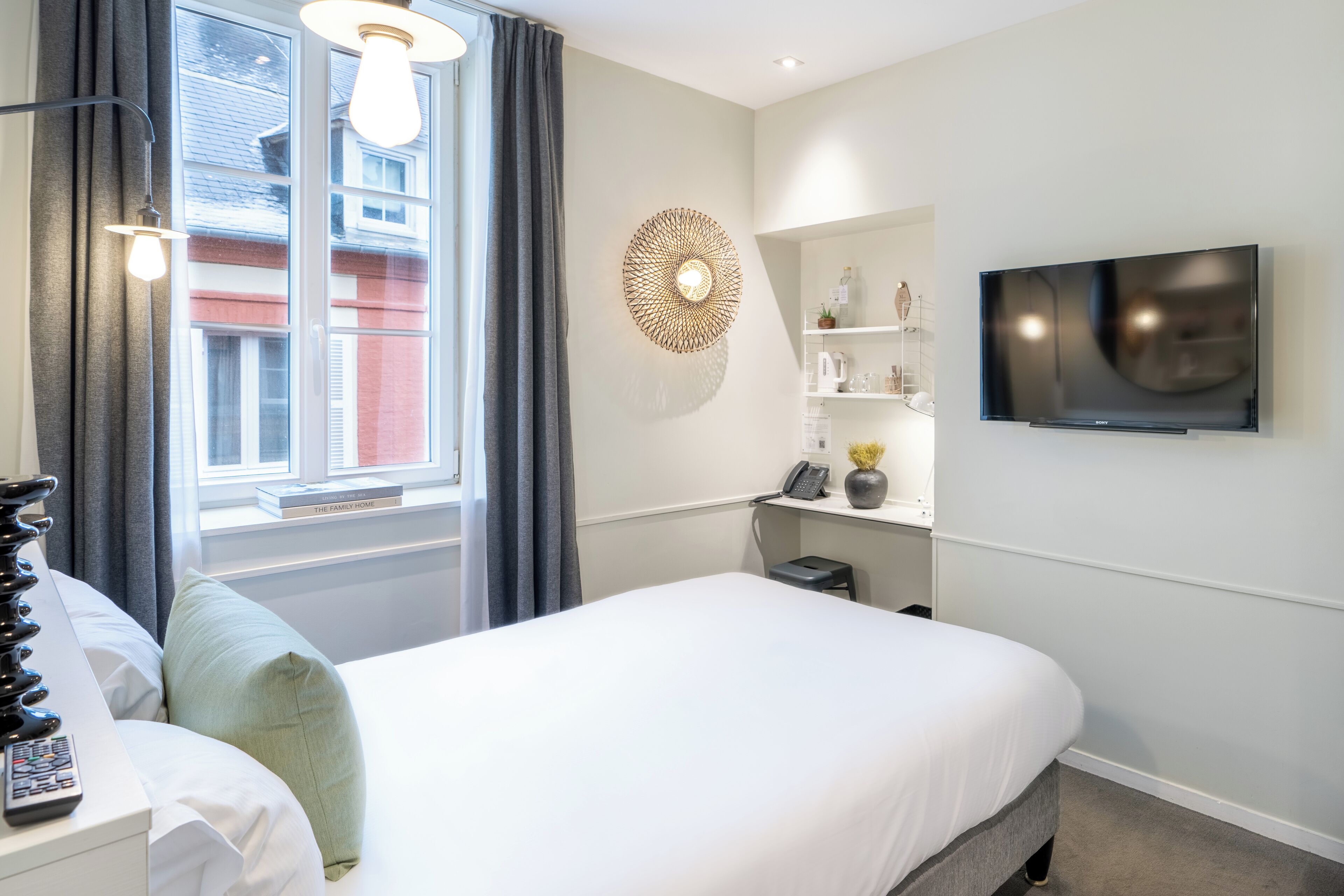Quarto de hotel moderno com uma cama grande, televisão na parede, mesa de cabeceira com abajur e janela com cortinas.