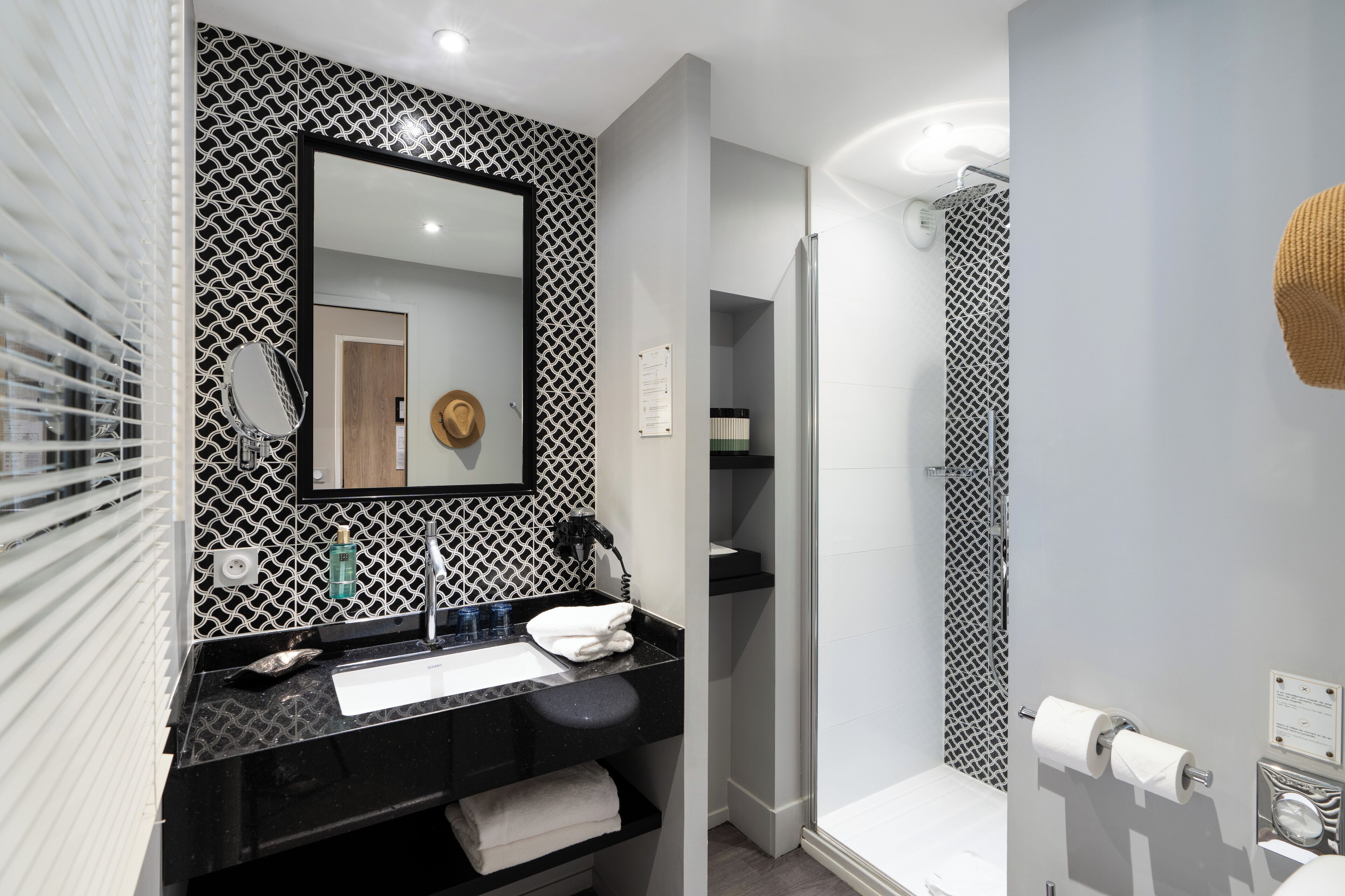 A modern bathroom with a black and white patterned wall, a large mirror, a sink with a towel, and a shower area.