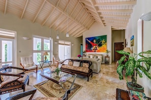 A spacious living room with a high ceiling, large windows, and a variety of seating options including a sofa, chairs, and a coffee table. - Sadewa Villa (Bequia Island)