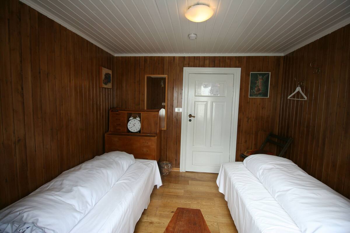 Una habitación con paneles de madera, dos camas individuales, una puerta blanca, un reloj y un pequeño espejo sobre un tocador.