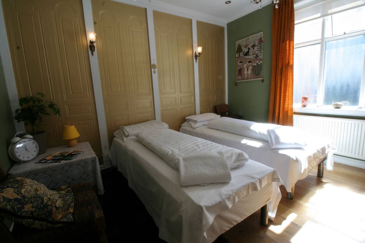 Habitación de hotel con dos camas, una mesita con lámpara, un reloj y una ventana con cortinas.