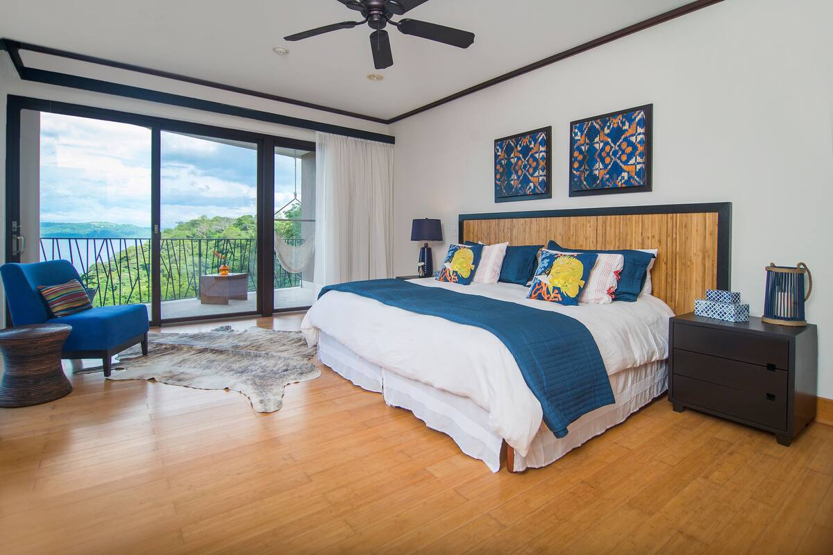 A bedroom with a large bed, a blue chair, a nightstand, and a view of the outdoors.