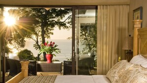 Quarto com uma grande porta de correr de vidro que dá para uma varanda com vista para o mar. Há uma cama com lençóis brancos e um vaso com planta vermelha na varanda.