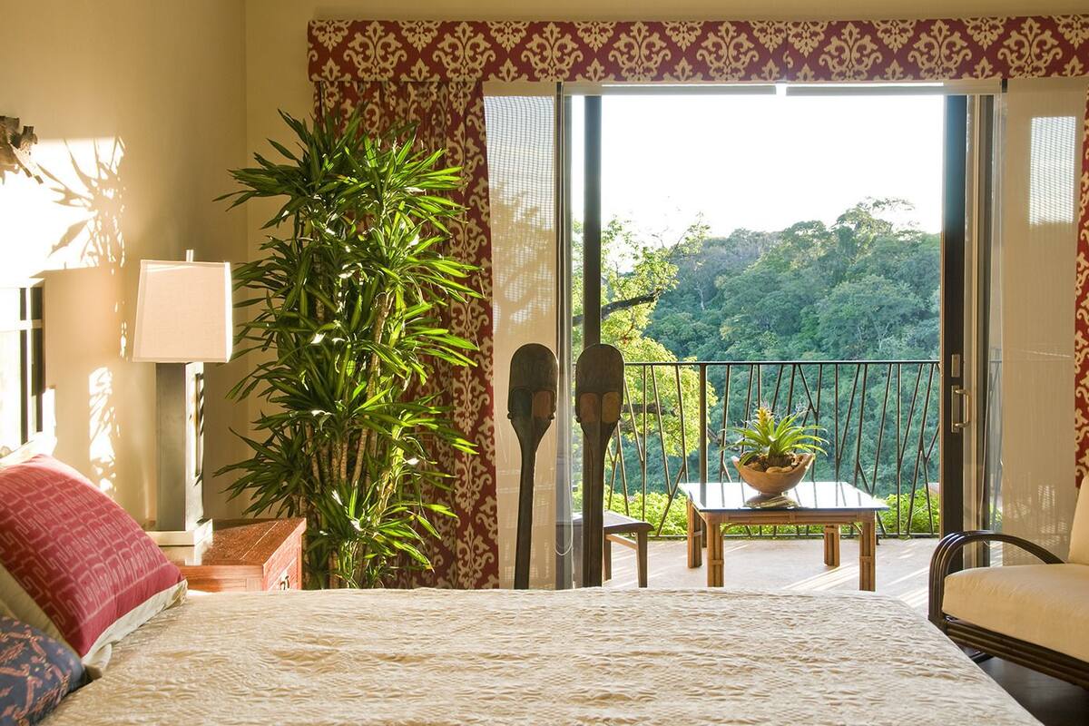 A hotel room with a bed, a table with a potted plant, a chair, a lamp, and a view of a balcony with a railing and greenery outside.