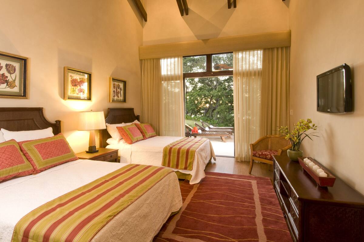 A hotel room with two beds, a large window with curtains, a flat-screen TV, and a wooden dresser.