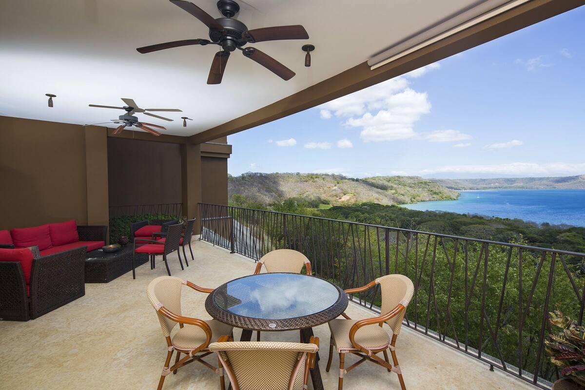 A balcony with a round table and chairs, a sofa with red cushions, and a ceiling fan.