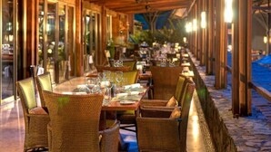 Restaurante ao ar livre com cadeiras e mesas de vime, copos de vidro e piso de pedra.