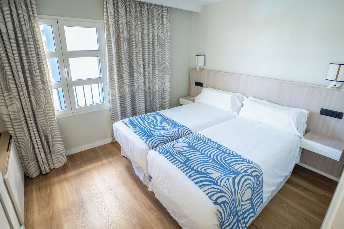 Una habitación de hotel con una cama grande, una mesita de noche, una lámpara de pared y una ventana con cortinas.