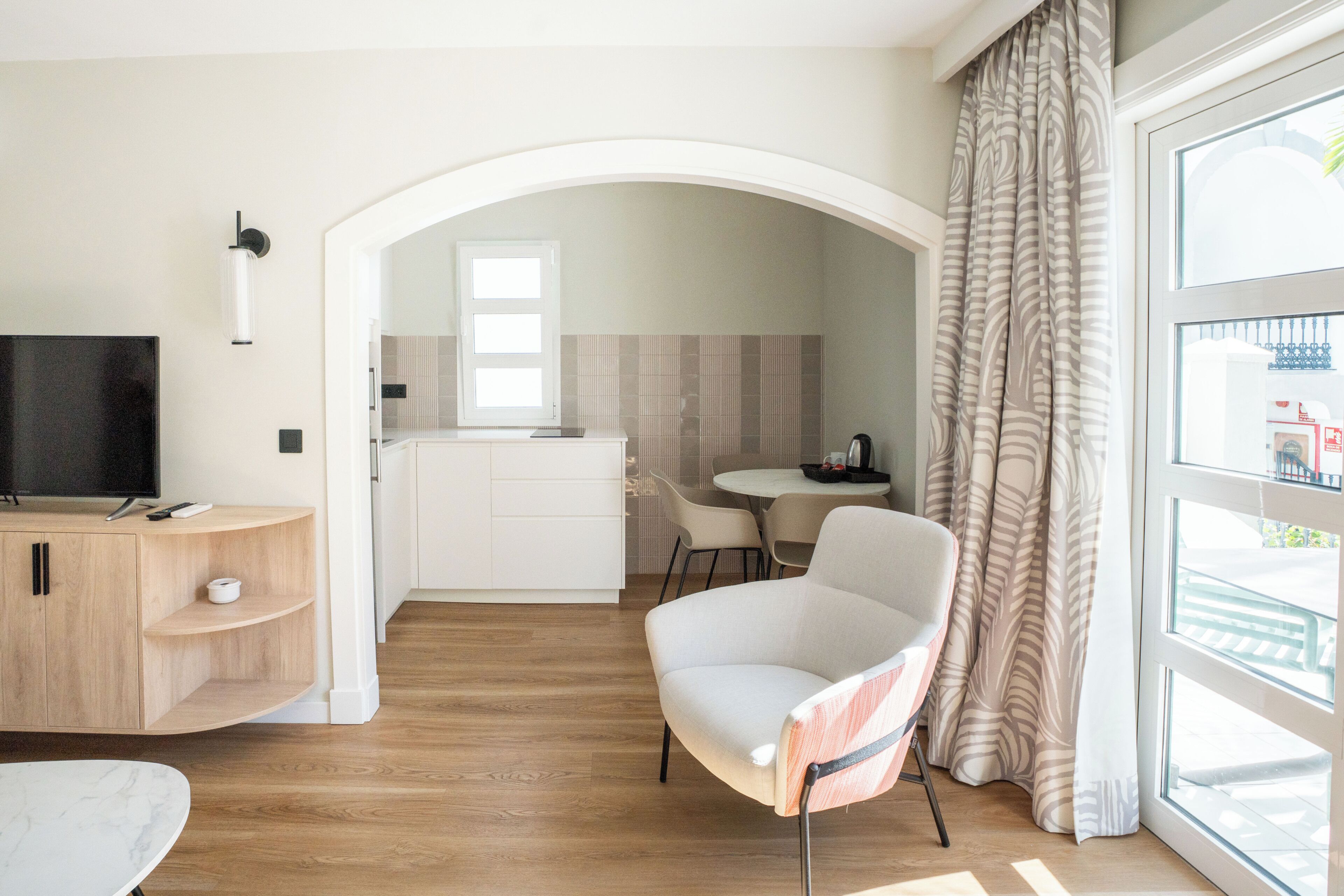 Een moderne hotelkamer met een televisie, een houten kast, een zithoek en een groot raam met gordijnen.