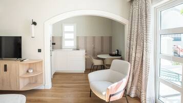 A modern hotel room with a TV, a wooden cabinet, a dining area, and a large window with curtains.