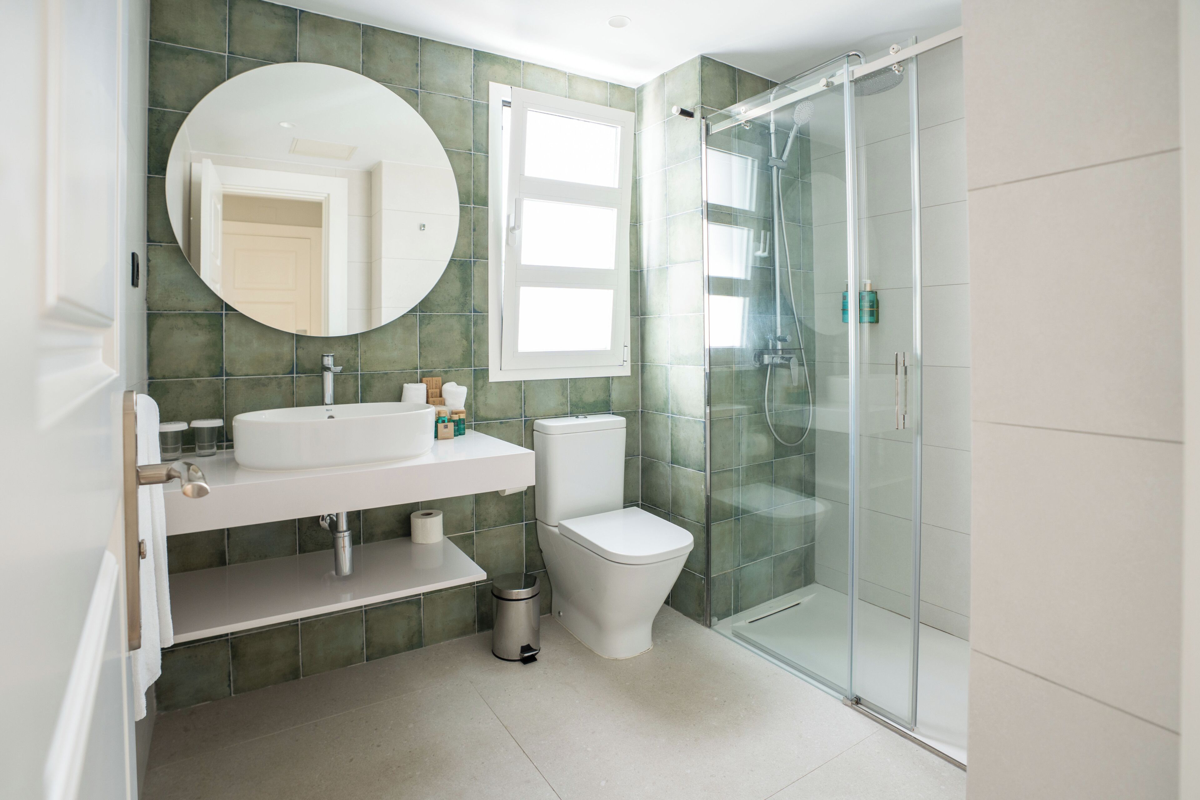 Un baño moderno con ducha de vidrio, sanitarios blancos y pared con azulejos verdes.