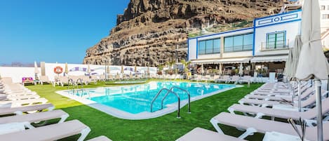 Zona de piscina con sillones reclinables, un bar y vistas a acantilados rocosos.